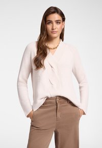 Femme aux longs cheveux bruns portant un pull côtelé rose clair et un pantalon marron, les mains dans les poches, avec un collier en chaîne dorée, sur un fond uni.