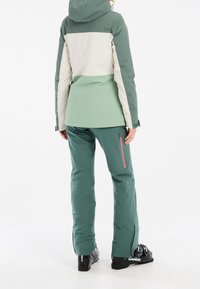 Ensemble veste et pantalon de neige en vert et crème. La veste est dotée d'une capuche et d’un design bicolore. Le pantalon possède une poche zippée et des guêtres aux chevilles.