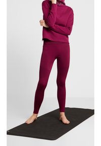 Sada aktivního oblečení ve vínové barvě zahrnující dlouhý rukáv s vysokým límcem a odpovídající legíny s texturovanými panely, vystavená na černé podložce.