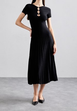 Kobieta w czarnej prążkowanej sukience midi z krótkimi rękawami i trzema ozdobnymi wycięciami na dekolcie, w parze z czarnymi szpiczastymi butami.