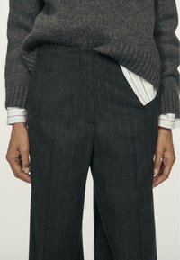 Dunkelgraue Wollhose mit einem Nadelstreifenmuster, entspannte Passform, flacher Front und Seitentaschen, kombiniert mit einem gestreiften weißen Hemd.