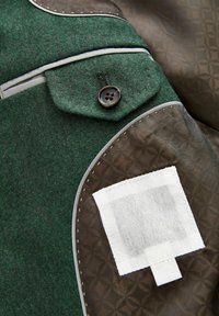 Jachetă verde din lână cu clapa buzunarului cu nasture și petic maro cu model pe cot, având un petic dreptunghiular alb cusut.