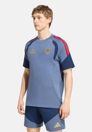 Jeune homme portant un vêtement de sport bleu avec des bandes rouges et des logos dorés, debout avec une expression neutre sur un fond uni.