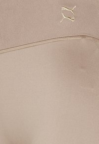 Leggings beige a vita alta realizzati in tessuto elastico, con una cintura a coste e un piccolo logo giallo sul lato. Texture complessivamente liscia.