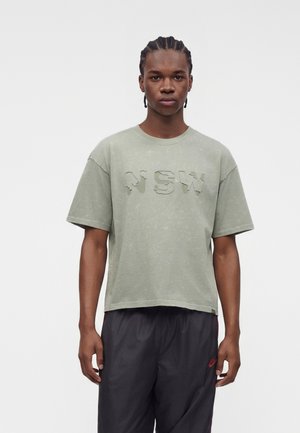 Jeune homme aux cheveux tressés portant un T-shirt vert délavé avec "NEW" en relief sur le devant et un short de sport noir sur un fond uni.