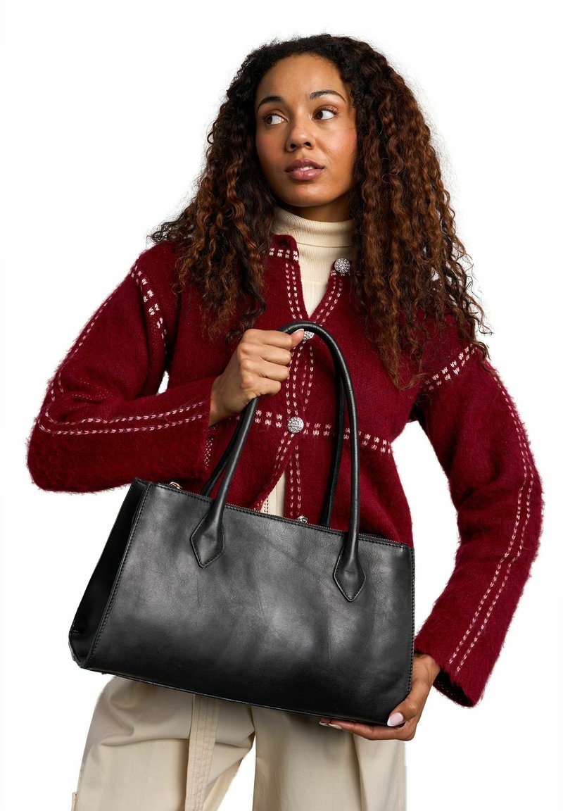Femme portant un cardigan rouge à motifs et un pantalon beige, tenant un grand sac cabas en cuir noir avec double anses.