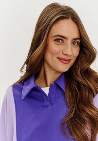 Fioletowy pulower z kołnierzykiem, wykonany z teksturowanej tkaniny z kolorowym blokowym wzorem. Miękki materiał podkreślający luźny krój.