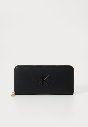 Portefeuille en cuir noir avec logo 'cK' en relief, fermeture éclair et quincaillerie dorée. Texture lisse et design rectangulaire.