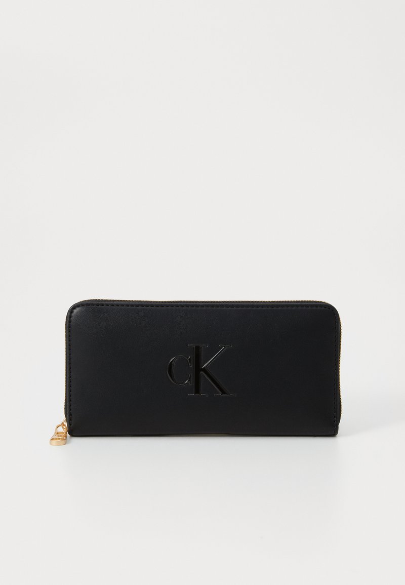 Portofel din piele neagră cu logo-ul 'cK' embosat, închidere cu fermoar și accesorii de culoare aurie. Textură netedă și design rectangular.