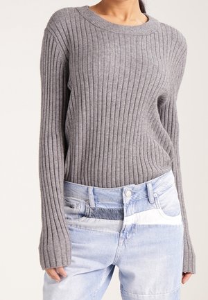 Vrouw draagt een grijze geribde trui met lange mouwen en tweekleurige lichtblauwe patchwork spijkerbroek, staand tegen een effen achtergrond.