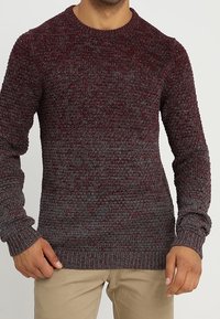 Pull en maille marron avec un motif texturé, col rond et manches longues. Ourlet inférieur et poignets côtelés. Étiquette logo notable sur le côté.