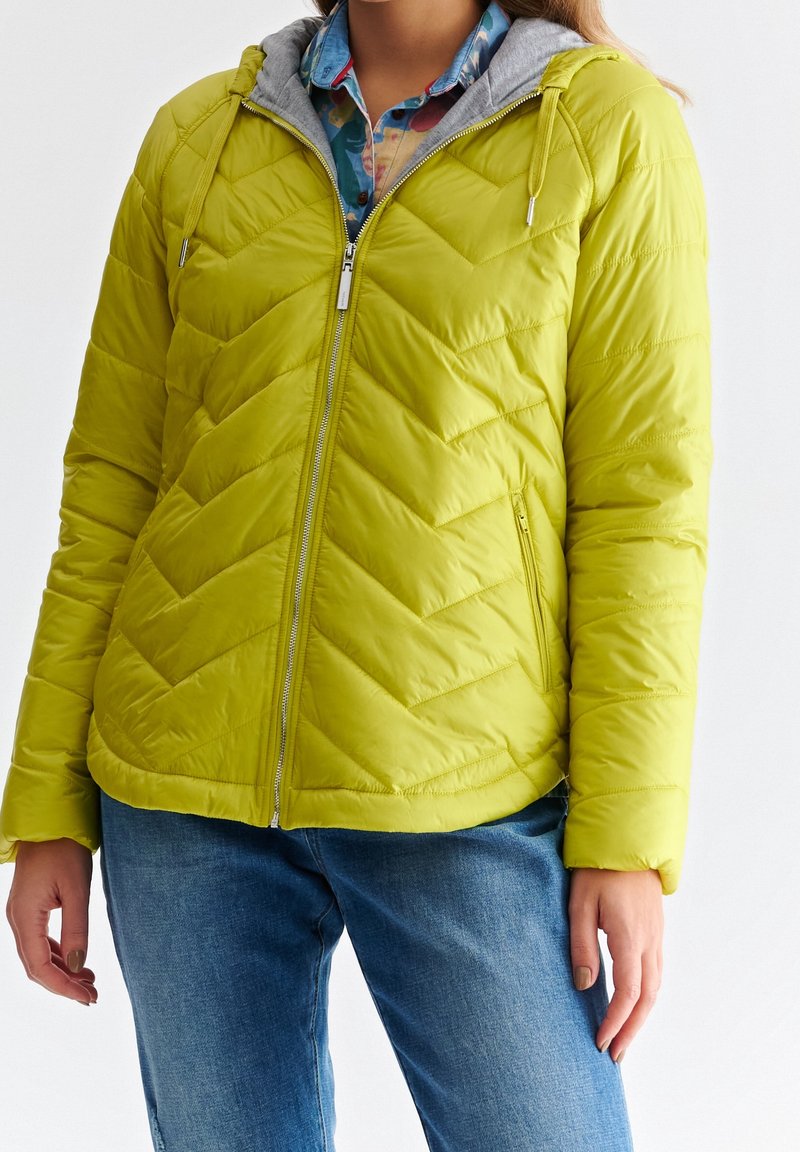 Personne portant une veste matelassée vert citron avec une fermeture éclair devant, par-dessus une chemise bleue à motifs et un jean bleu, debout devant un fond blanc.