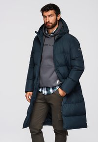 Donkergroene lange gewatteerde jas met een ritssluiting en een capuchon. Hieronder zijn een grijze sweatshirt en een geruit overhemd zichtbaar.