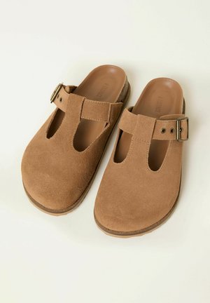 Ruskeat mokkanahkaiset liukusäädettävät puukengät, joissa on leikatut kuviot ja säädettävä hihna. Varustettu pehmustetulla sisäpohjalla ja kuvioidulla kumipohjalla.