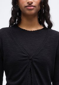 Μαύρο ριμπ πλεκτό ζακέτα με V-λαιμόκοψη, κυματιστή άκρη και κουμπιά στο εμπρός μέρος. Διαθέτει υφή στο ύφασμα και εφαρμοστή σιλουέτα.