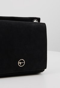 Borsa a tracolla nera in finta pelle scamosciata con una superficie liscia, forma rettangolare e un accentuata logo circolare in argento sulla parte anteriore.