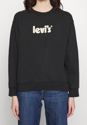 Personne portant un sweat-shirt Levi's noir avec un logo blanc sur la poitrine et un jean bleu, debout devant un fond clair uni.