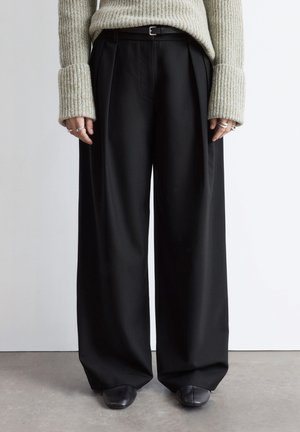 Black wide-leg trousers with pleats, made from a smooth fabric. Paired with a long, ribbed, light-colored sweater and black shoes.