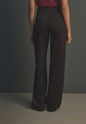 Personne portant un pantalon large noir à fines rayures, taille haute, debout les bras détendus le long du corps sur un fond neutre.