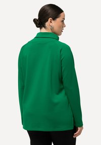 Top verde a maniche lunghe con colletto alto, caratterizzato da una superficie liscia e spacchi laterali per maggiore comodità. Nessun motivo o accentuazione visibile.