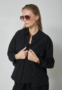 Melnā pogājām uzvilkta krekla ar divām priekšējām kabatām, valkāta pār saskaņotu melnu topu. Modele valkā lielas rozā toni lēcošas saulesbrilles ar zelta rāmjiem.