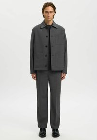 Graham gewebte Jacke mit Kragen, Frontknöpfen und zwei aufgesetzten Taschen, kombiniert mit passenden grauen Hosen und schwarzen Schuhen.