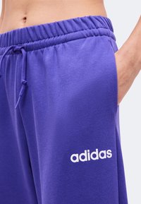 Κοντινό πλάνο σε μωβ παντελόνια φόρμας Adidas με ελαστική μέση και κορδόνι, που δείχνει ένα χέρι στην τσέπη και ένα μέρος γυμνής κοιλιάς.