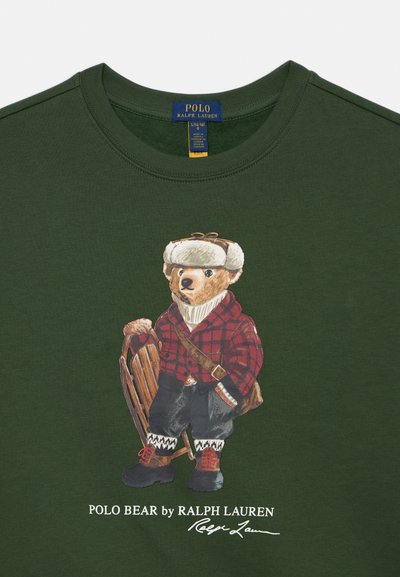 Zelené bavlnené tričko s ilustráciou medveďa v červenej kockovanej košeli, ktorý drží drevený sanie. Obsahuje biely potlačený text pod grafikou.