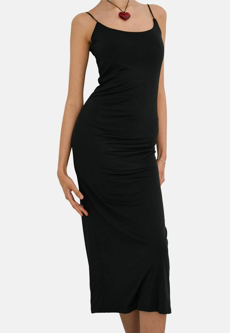 Femme portant une robe midi noire ajustée avec de fines bretelles et un collier pendentif en forme de cœur rouge sur un fond uni.