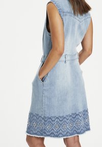 Hihaton vaaleansininen farkkumekko, jossa taskut ja koristeellinen tikkireuna timanttikuvioilla, päällä henkilö, joka on kuvattu takaa.