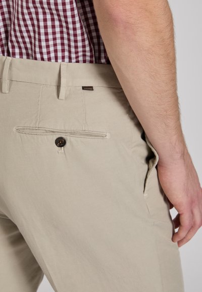 Beżowe spodnie z tylną kieszenią zapinaną na ciemny guzik, na tle koszuli w kratę w kolorze bordowo-białym.