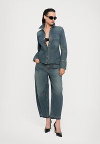 Jeansjacke und weiter Jeans in einer dunklen Waschung, mit genähten Details, Knopfverschlüssen und einem lässigen Schnitt. Kombiniert mit schwarzen High-Heels.