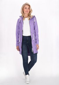 Lys lilla vandtætte jakke med snorehue, frontlynlås og hvide detaljer. Båret over en hvid top og mørke jeans.