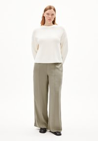 Weißer strukturierter Pullover mit weiten Ärmeln, kombiniert mit olivgrünen weit geschnittenen Hosen. Das Model trägt schwarze Schuhe und steht vor einem weißen Hintergrund.