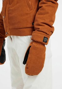 Bruine corduroy jas met ribbelstructuur, voorzien van een v-hals en grote knopen. Gecombineerd met oversized bruine wanten en witte broeken.