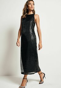 Robe maxi sans manches noire avec un motif de sequins en grille, présentant une silhouette droite et des détails de fente sur les côtés. Associée à des escarpins à lanières.