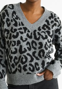 Grauer Strickpullover mit V-Ausschnitt, der ein schwarzes Leopardenmuster aufweist. Gerippte Bündchen und Saum verleihen Textur und Detail.