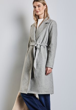 Manteau en laine gris clair avec une ceinture, un col châle et des manches longues. Porté sur un haut blanc et associé à un jean large bleu foncé.
