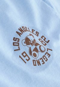 Vaaleansininen puuvillakangas, jossa on ruskea painatus nallekarhusta, tekstillä "LOS ANGELES LEGEND 1982". Sujuva rakenne.