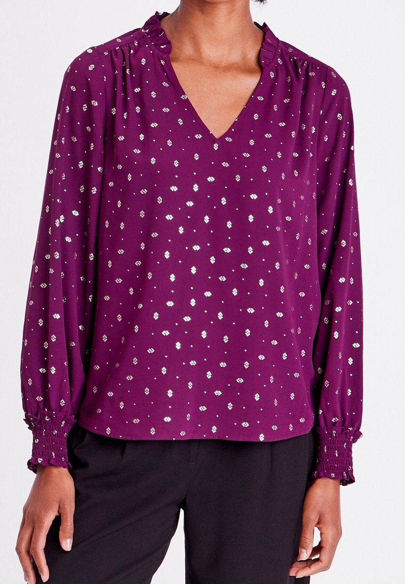 Blouse violette avec un décolleté en V, un col à volants et des manches longues. Elle présente un motif floral argenté et des poignets froncés pour un look texturé.