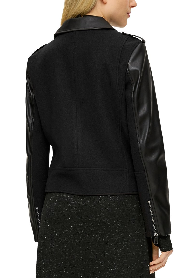 Schwarze Jacke mit Wollkörper und Lederärmel, kurz geschnitten, mit flachem Kragen und seitlichen Reißverschlussakzenten. Insgesamt glatte Textur.