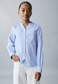 O tânăr cu părul închis la culoare prins la spate, îmbrăcat cu o cămașă cu mânecă lungă, albă cu dungi albastre, și pantaloni albi, stând cu o mână în buzunar.