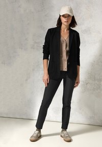 Černý blazer přes béžovou košili na knoflíky, v kombinaci s černými kalhotami a béžovými teniskami. Model má na sobě čepici světlé barvy. Hladké, přiléhavé látky.