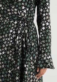 Schwarzes Wickelkleid mit einem Sterne-Muster in Grün, Weiß und Grau. Lange Ärmel mit Rüschensäumen und einem Bindeband in der Taille.