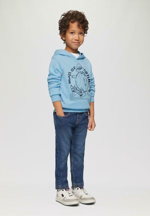 Hoodie bleu avec un motif graphique, jean en denim long, et baskets grises. Cheveux bouclés, mains dans les poches, se tenant avec un léger sourire.