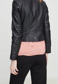 Kvinna i en figursydd svart läderjacka över en persikofärgad blus och svarta byxor, sedd bakifrån mot en enfärgad bakgrund.