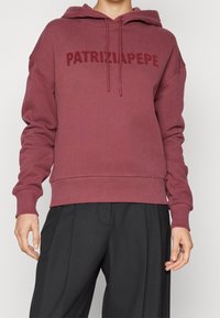 Felpa con cappuccio color bordeaux con un logo frontale che recita "PATRIZIA PEPE", caratterizzata da una vestibilità comoda, polsini e orlo a costine. Tessuto morbido.