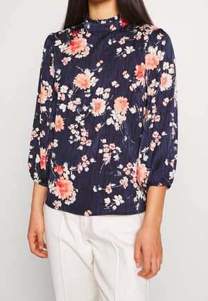 Kobieta nosząca granatową bluzkę z wysokim kołnierzem, różowo-białym kwiatowym wzorem i długimi bufkowatymi rękawami, zestawioną z białymi spodniami.