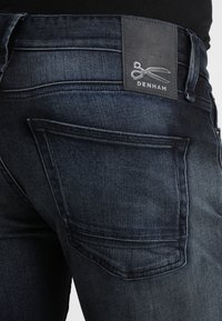 Mörkblå denimjeans med en något blekt finish, med en läderlapp som har "Denham"-logotypen och en bild av en sax på baksidan.