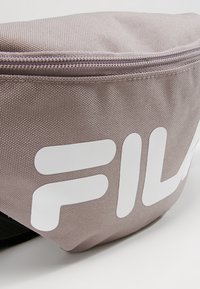 Pochette en tissu texturé marron clair avec une fermeture éclair et un grand logo partiel blanc "FILA" imprimé sur le devant.